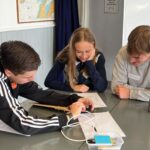 Image of three students working collaboratively at a table, paper and cables on table. They are smiliing.